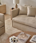 A cozy living room featuring the handmade-in-Los Angeles Dume Sofa in beige, an armchair, a wooden side table with a lamp and cup, a round coffee table with books and white flowers, and a light-colored rug on wooden floors.