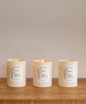 Three white Hudson Glass Candles, each with a printed jenni kayne label, are lit and arranged side by side on a wooden surface, highlighting the hand-poured Los Angeles natural candle against a simple, light background.