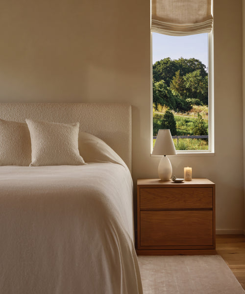 A cozy bedroom features a bed with white linens and pillow, a wooden nightstand with a white lamp and the Hudson Glass Candle. Sunlight pours through the window, revealing green trees and fields outside.