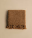 The Hudson Throw, a folded brown fuzzy alpaca blanket with fringe along the bottom edge, rests on a plain beige background.