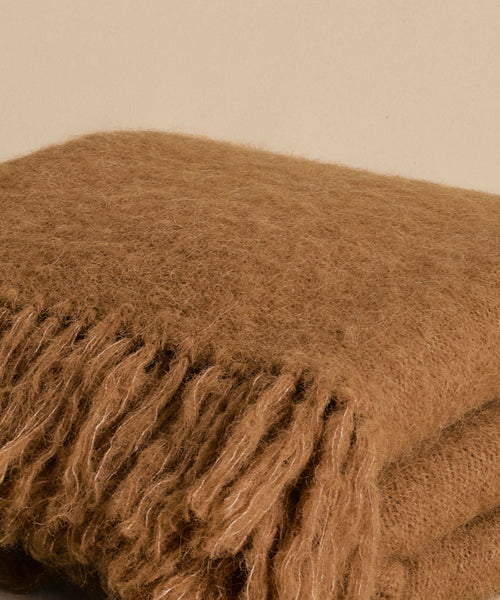 A close-up of the Hudson Throw, featuring a soft, fuzzy texture and long fringes, folded against a plain beige background—a cozy, lightweight blanket ideal for any setting.
