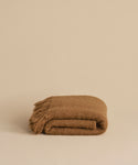 The Hudson Throw, a folded brown alpaca blanket with fringed edges, is displayed on a plain beige surface against a matching beige background.