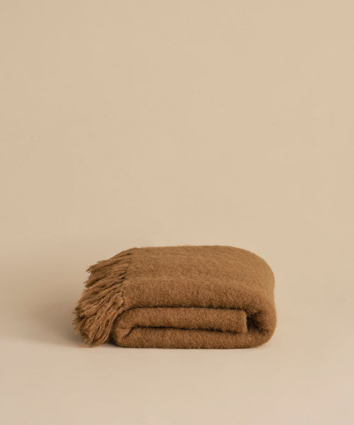 The Hudson Throw, a folded brown alpaca blanket with fringed edges, is displayed on a plain beige surface against a matching beige background.