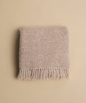 The Hudson Throw, a soft beige alpaca scarf with fringed edges, is neatly folded on a matching beige surface.
