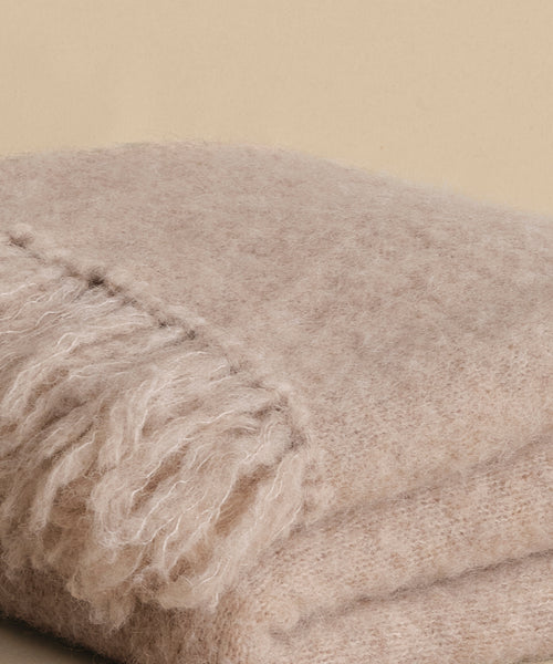 A close-up of the Hudson Throw, a soft, light beige alpaca blanket with fringed edges, folded neatly on a neutral background.