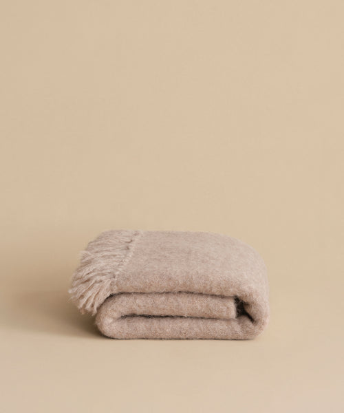 The Hudson Throw, a folded beige alpaca blanket with fringe on one end, is displayed on a neutral beige background.