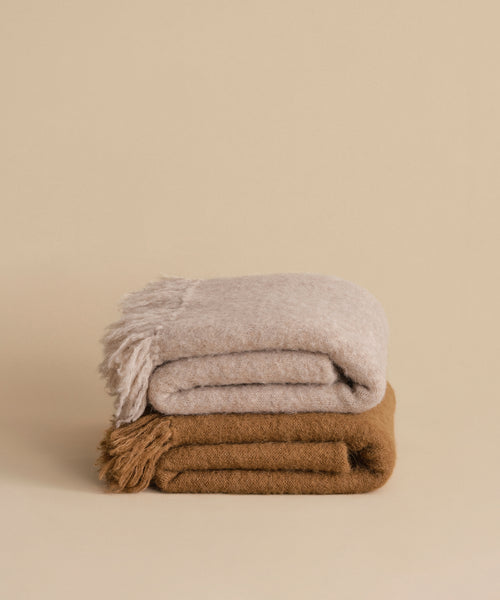 Two neatly folded Hudson Throw blankets are stacked against a beige background; the top is a light beige lightweight throw with fringed edges, and the bottom is solid brown. Perfect for adding cozy Hudson Valley style to your space.