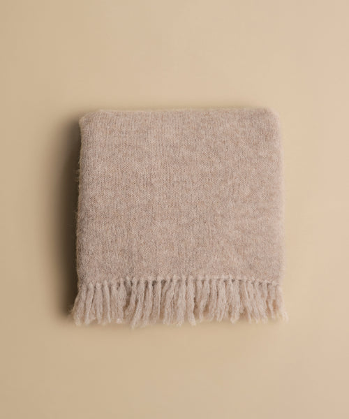 The Hudson Throw, a soft beige alpaca scarf with fringed edges, is neatly folded on a matching beige surface.