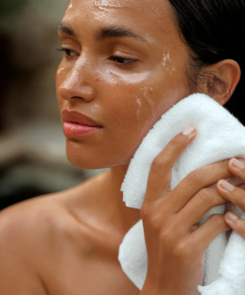 After using the Hydrating Double Cleanse Duo, a woman with dewy skin gently presses a white towel to her cheek, looking relaxed and refreshed against a soft natural background.