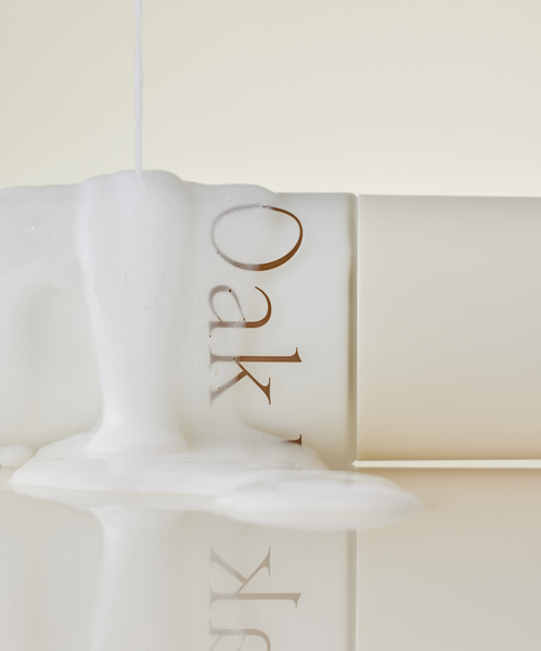 A creamy, moisture-rich cleanser from the Hydrating Double Cleanse Duo pours over a light Oak-labeled bottle, creating foamy overflow on a glossy surface. The softly lit, minimal scene reflects gentle care for your skin barrier.