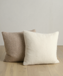 Two textured square pillows rest on a light wooden floor against a pale wall: one is the beige Alpaca Basketweave Pillow, and the other is an off-white super soft pillow.