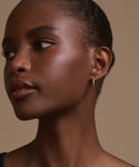 A close-up of a person with dark skin, natural makeup, and short hair pulled back, wearing the Iris Diamond Hoops in 14k gold, looking to the left against a neutral beige background.