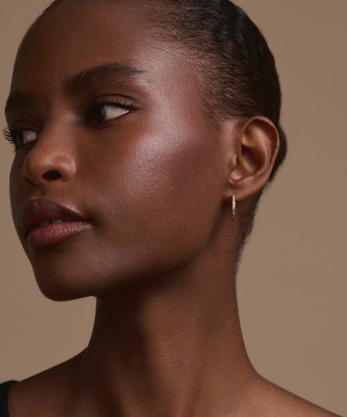A close-up of a person with dark skin, natural makeup, and short hair pulled back, wearing the Iris Diamond Hoops in 14k gold, looking to the left against a neutral beige background.