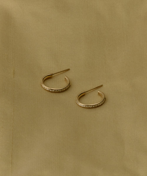 The Iris Diamond Hoops are small 14k gold earrings adorned with sparkling stones, elegantly displayed on a light beige fabric—ideal for anyone seeking timeless heirloom jewelry.