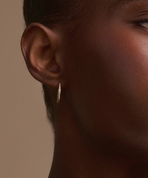 A close-up of a person’s ear and face, showcasing the elegant Iris Diamond Hoops—14k gold earrings with small embedded stones—set against a neutral beige background.