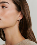 Close-up of a woman’s face in profile, featuring smooth skin and straight brown hair. She wears the Iris Diamond Hoops—gold earrings with embedded stones. The neutral background is soft and out of focus.
