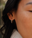 Close-up of a womans ear wearing Jenni Kaynes Iris Diamond Hoops—14k gold earrings with small diamonds. Her eye is closed as her dark hair softly frames her face against a blurred background.