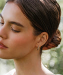 A woman with fair skin and brown hair in a low bun stands outdoors with her eyes closed, wearing delicate Iris Diamond Hoops in 14k gold. Blurred greenery serves as the background.