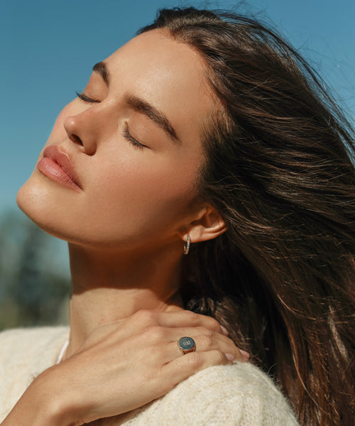A woman with long brown hair stands outdoors, eyes closed and enjoying the sunlight. She wears a light sweater, the Iris Diamond Hoops by Jenni Kayne, and a ring, with her hand resting gently on her shoulder against a blue sky.