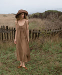 Woman in a brown dress and hat standing in a field with a wooden fence in the background