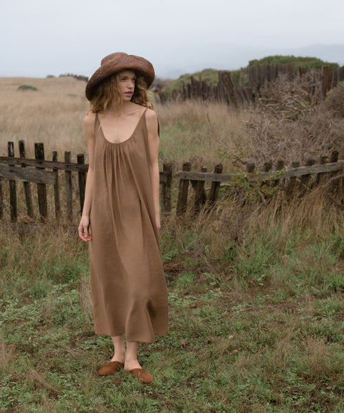 Woman in a brown dress and hat standing in a field with a wooden fence in the background
