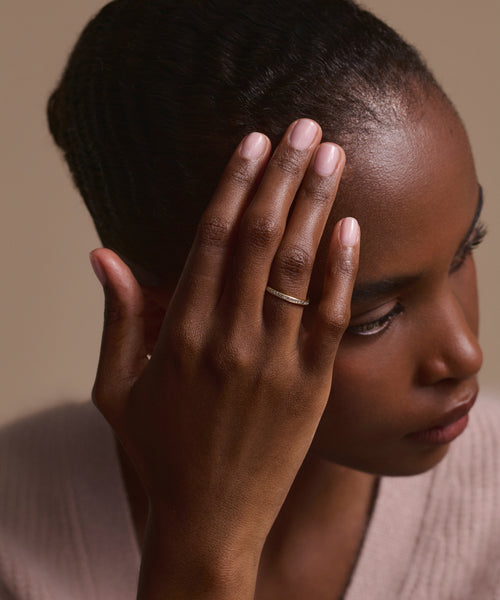 A woman with smooth skin and manicured nails touches her face, showcasing the Iris Eternity Band in 14k solid gold with conflict-free diamonds, set against a beige background and wearing a soft neutral top.