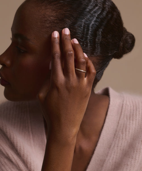 A woman in a soft pink sweater touches her ear, showcasing the elegant Iris Eternity Band in 14k solid gold. Her manicured pale pink nails add to her sophisticated look.