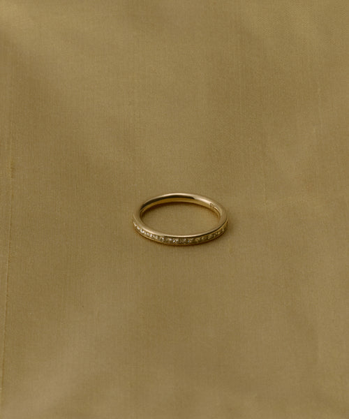 The Iris Eternity Band, a slim 14k solid gold ring set with small, conflict-free diamonds, rests on a plain light brown fabric background.