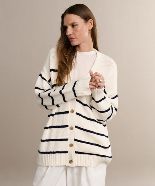 A woman with long brown hair models the Chloe Cardigan in Ivory Navy Stripe over a white top and pants, standing against a neutral background with a relaxed expression.