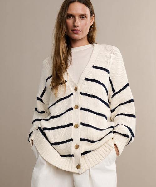 A woman models the Chloe Cardigan in ivory navy stripe with wooden buttons over a white top, hands in white pants pockets, highlighting this stylish sweater against a plain beige background.