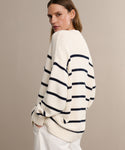 A person wearing the Chloe Cardigan in Ivory Navy Stripe with white pants stands against a plain beige background, looking over their shoulder at the camera.