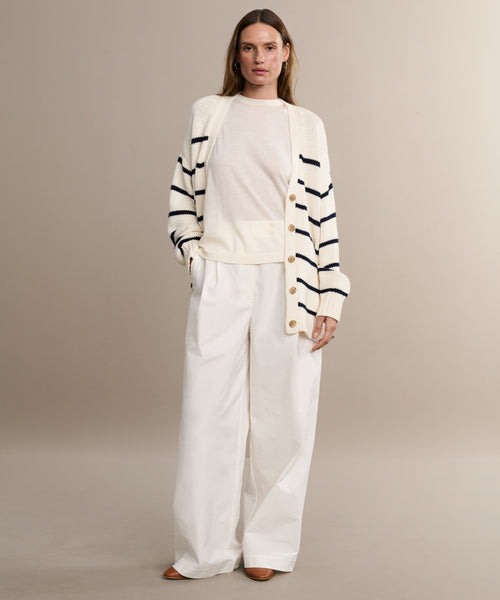 A woman stands against a neutral background wearing the Chloe Cardigan in Ivory Navy Stripe over a white top, paired with wide-leg white trousers and brown shoes. Her hands are in her pockets, offering a chic warm-weather sweater look.