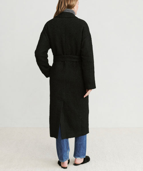 A person with shoulder-length hair stands facing away, wearing the Jacquard Overcoat belted over blue jeans and black slip-on shoes. The background is plain and light-colored.