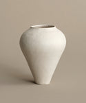 The JW2 Vase is a minimalist, handcrafted France stoneware piece with a wide, rounded body and narrow, tapered base. Its slightly textured surface and elegant matte off-white finish stand out against a plain beige background.
