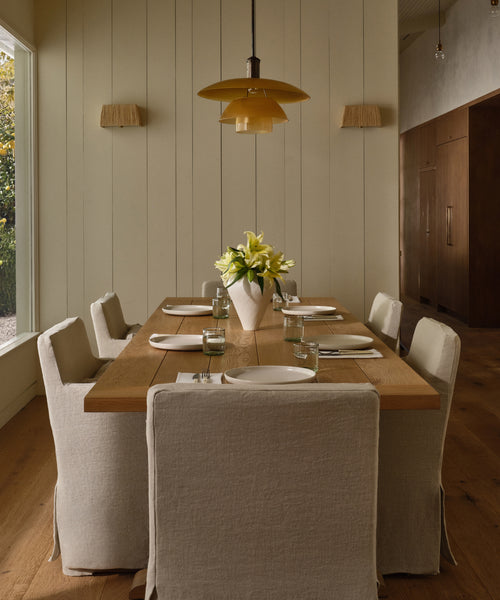 A modern dining room features a wooden table set for six with beige slipcovered chairs. The centerpiece is the matte white JW2 Vase filled with white lilies. A yellow pendant light hangs above, while sunlight streams in from the left window.