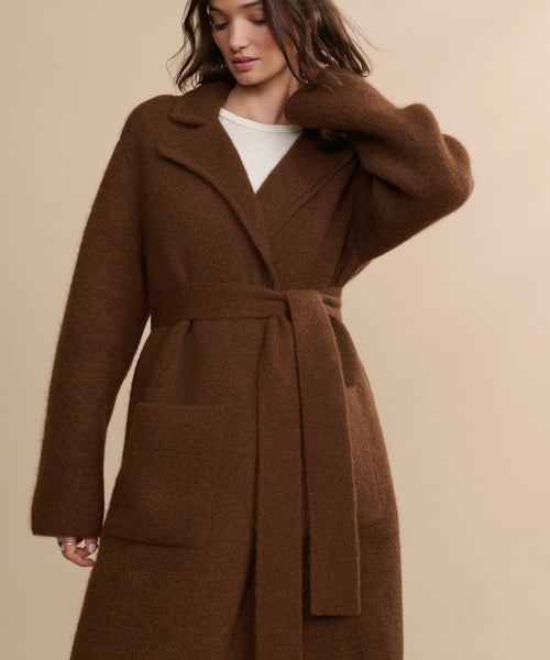 A woman stands against a beige background, wearing the Jessica Sweater Coat—a long, brown, alpaca blend coat—over a white top. Her hair is loose and she raises one hand to her head.