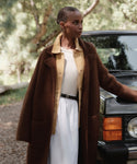A woman stands outdoors by a black car, wearing the Jessica Sweater Coat—a long brown alpaca blend—over a beige shirt, cream top, and white skirt with a black belt. She gazes to the side, with trees in the background.