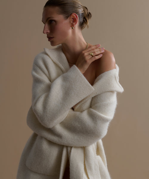 A woman with short, slicked-back hair models the Jessica Sweater Coat in ivory, paired with minimal gold jewelry. She stands against a neutral background with arms crossed, gazing thoughtfully to the side.