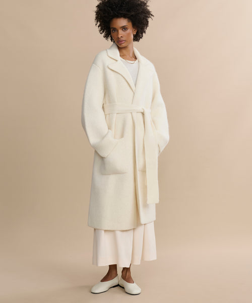 A woman with short, curly hair stands against a beige background wearing the Jessica Sweater Coat, an ivory alpaca blend, over a cream outfit and white shoes. Her hands are in the coat pockets.