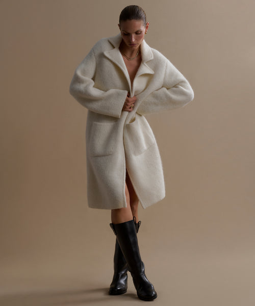 Against a beige backdrop, a person wears the Jessica Sweater Coat—an oversized, long ivory alpaca-blend piece—paired with black knee-high boots. Their head is slightly bowed as they adjust the front of the coat.