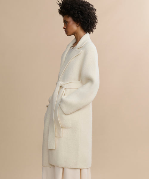 A woman with curly hair wears the ivory Jessica Sweater Coat, which is belted and made from an alpaca blend, paired with light pants, standing in profile against a beige background.