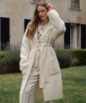 A woman with long wavy hair stands in a garden, wearing the elegant Jessica Sweater Coat—an ivory, long alpaca blend layer—over light-colored clothing, with a building and lush greenery behind her.