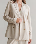 A woman models the Jones Blazer, a cream-colored double-breasted suit with matching pants and a relaxed, elegant fit, holding one side near the buttons against a plain background.