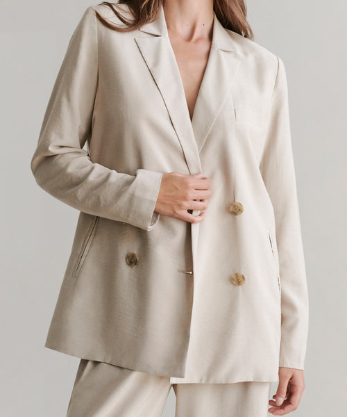 A woman models the Jones Blazer, a cream-colored double-breasted suit with matching pants and a relaxed, elegant fit, holding one side near the buttons against a plain background.