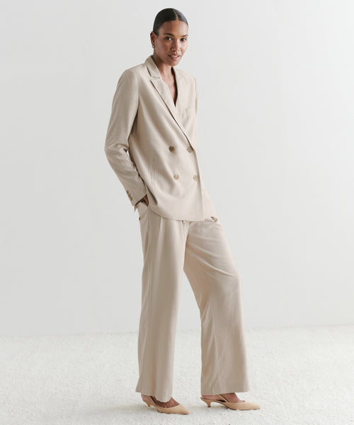 Wearing the Jones Blazer—a light beige, double-breasted lightweight blazer with wide-leg trousers and beige heels—a person stands smiling against a plain white background, hands in their pockets.