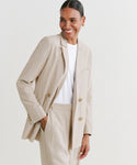 A woman smiles, wearing the Jones Blazer in light beige over a white shirt, with one hand in her pocket, standing against a plain white background.