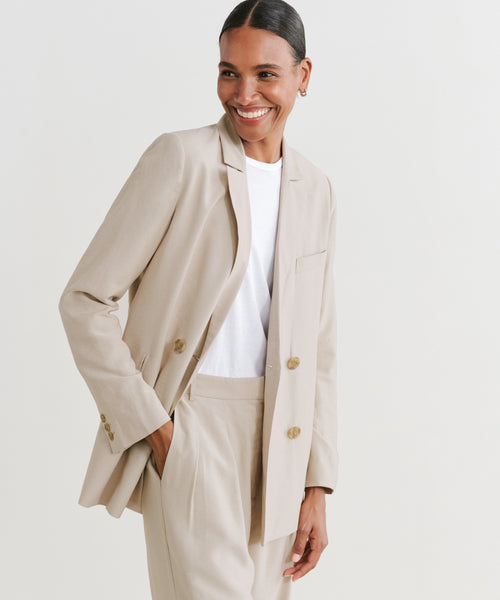 A woman smiles, wearing the Jones Blazer in light beige over a white shirt, with one hand in her pocket, standing against a plain white background.