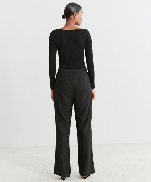 A woman faces away in a fitted black long-sleeve top and the Jones Trouser, featuring soft tailoring. Her hair is pulled back in a neat bun as she stands on a light floor against a plain white background.