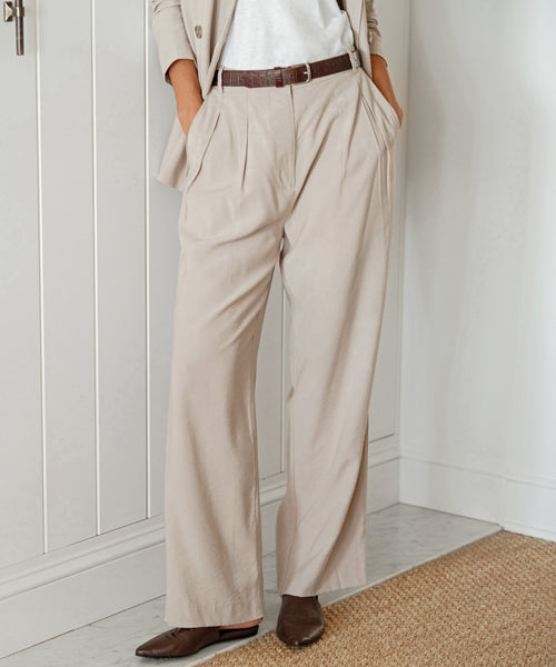 Indoors, a person wears high-waisted, wide-leg Jones Trouser with a matching blazer, brown belt, and pointed-toe shoes. Hands in pockets, they embody a modern power suit look. Their head is not shown.
