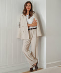 A woman stands against a white paneled wall in a modern beige power suit—featuring the Jones Trouser, matching blazer, and a white t-shirt. Her arms are crossed and her long brown hair is loose as she looks confidently at the camera.